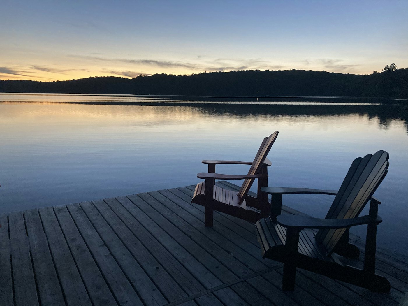 Two wooden Adirondack chairs on a dock facing a calm lake at sunset